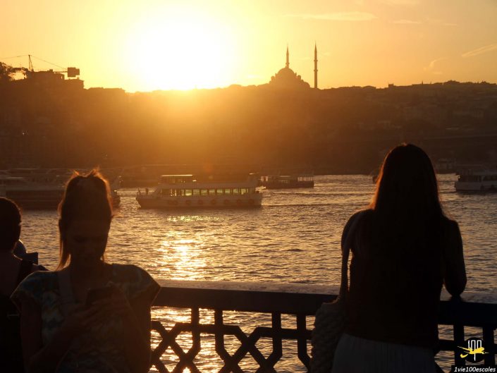 Deux personnes se tiennent sur un front de mer au coucher du soleil à Istanbul en Turquie, avec des bateaux sur l'eau et une mosquée avec deux minarets visibles en arrière-plan. Une personne regarde un smartphone tandis que l’autre regarde vers l’eau.