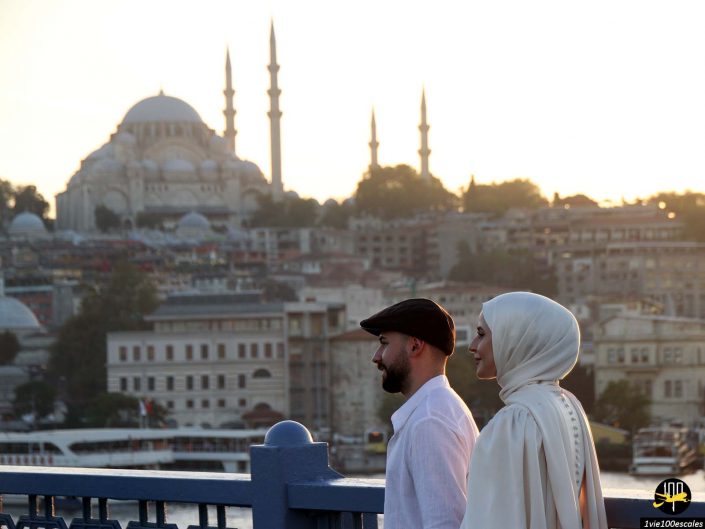 Un homme et une femme marchent côte à côte sur un pont au coucher du soleil, avec en arrière-plan un paysage urbain comportant une mosquée aux multiples minarets, à Istanbul en Turquie.