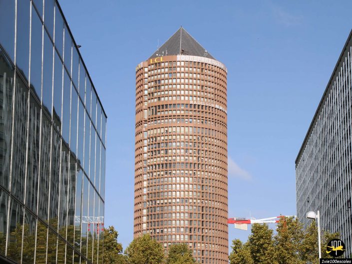 Un immeuble cylindrique de grande hauteur avec un toit triangulaire se dresse entre deux bâtiments à façade de verre de chaque côté et des arbres au premier plan, à Lyon en France.