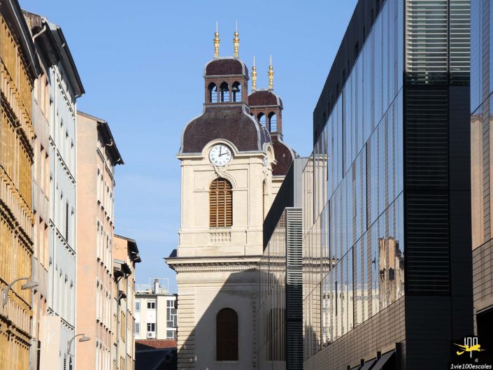 Une tour d'horloge aux dômes marron se dresse entre les bâtiments historiques et modernes, se reflétant dans une façade de verre voisine sous un ciel bleu clair, à Lyon en France.