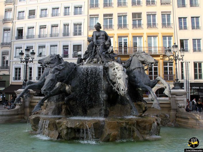 L'image montre la Fontaine Bartholdi à Lyon en France, avec une sculpture représentant une femme et quatre chevaux entourés d'eau, sur fond de bâtiments à plusieurs étages.