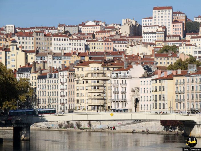 Un pont enjambe une rivière devant de nombreux immeubles à plusieurs étages aux toits de tuiles rouges sur une colline sous un ciel bleu clair, à Lyon en France.