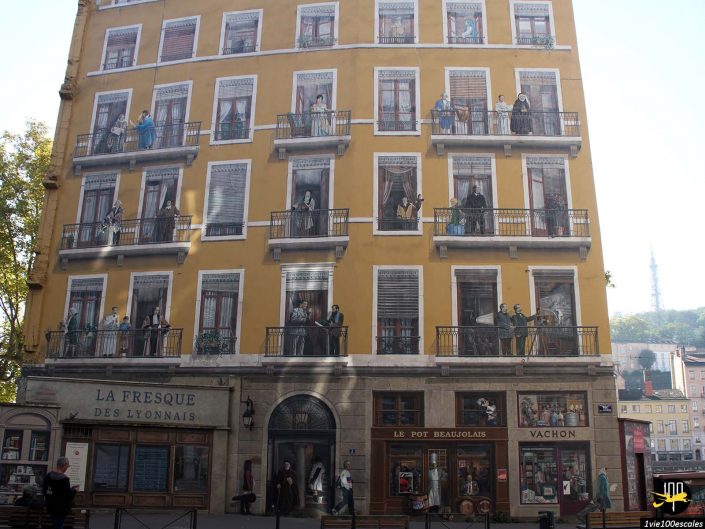 Une façade d'immeuble de cinq étages à Lyon en France avec des peintures murales réalistes représentant des personnes sur des balcons et des devantures de magasins, se fondant parfaitement dans l'environnement réel.