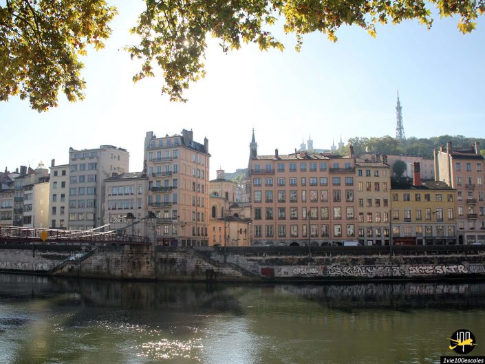 Une rangée de bâtiments historiques borde une berge sous un ciel clair à Lyon en France. Des graffitis recouvrent la digue de la rivière et une haute tour en acier se dresse sur une colline en arrière-plan.