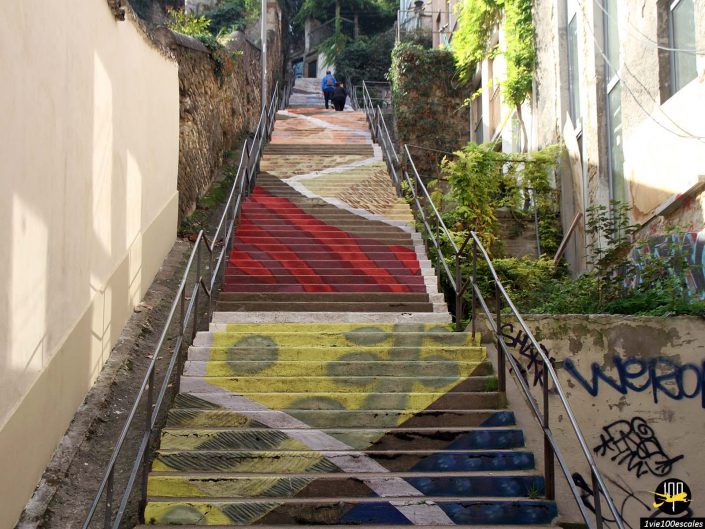 Un escalier multicolore avec des motifs peints vibrants monte entre les bâtiments à Lyon en France. Des graffitis décorent les murs et les marches. Deux personnes sont visibles en haut des escaliers.