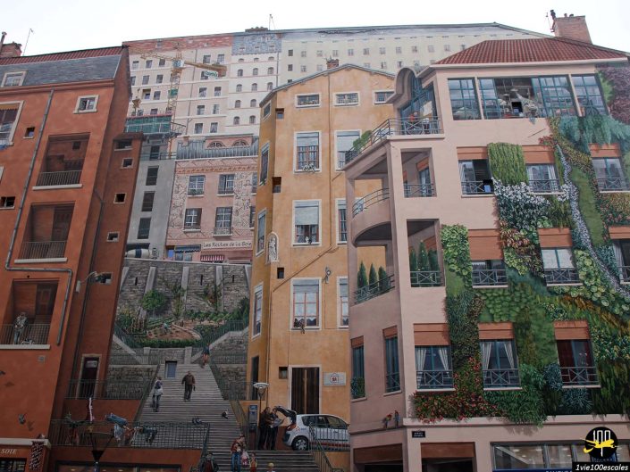 Une scène de rue à Lyon en France présente une fresque détaillée sur les façades des bâtiments. L'œuvre représente des bâtiments, des balcons, des fenêtres, de la verdure et des escaliers, créant l'illusion vivante d'un environnement urbain animé.