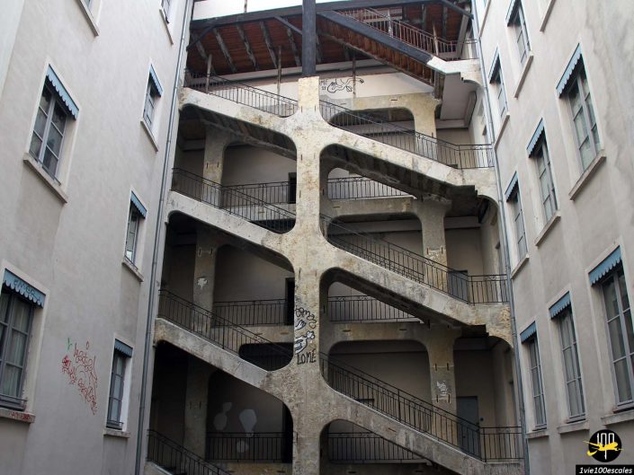 Une structure d'escalier apparente de plusieurs étages avec quatre niveaux, béton patiné et garde-corps, située entre deux bâtiments beige pâle d'aspect résidentiel à Lyon en France, avec des graffitis sur les murs et les surfaces d'escalier.