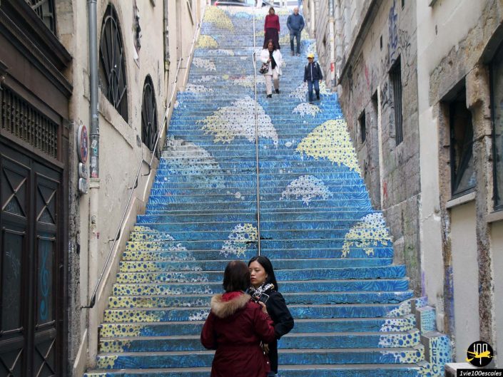 Des gens qui montent et descendent un escalier extérieur décoré avec une fresque murale bleue et jaune sur le thème de l'océan à Lyon en France. Deux femmes sont debout et discutent en bas des escaliers.