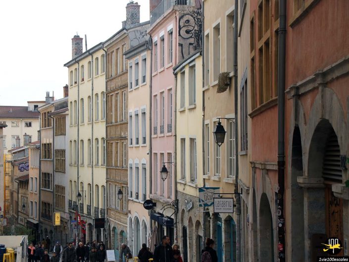 Gens marchant le long d'une rue bordée de bâtiments colorés à plusieurs étages dans une ville européenne à Lyon en France. Des panneaux et des lampadaires sont visibles et le ciel est couvert.