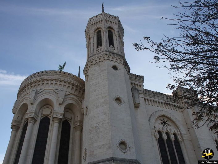 Une église en pierre blanche avec une tour cylindrique ornée et des fenêtres cintrées se dresse majestueusement à Lyon en France, avec une branche d'arbre au premier plan sous un ciel bleu immaculé.