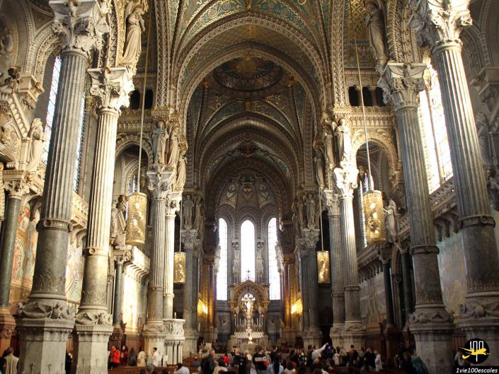 Intérieur d'une grande cathédrale à Lyon en France avec un plafond et des colonnes richement décorés. L'allée centrale mène à un autel orné, éclairé par la lumière naturelle provenant de grandes fenêtres cintrées. Les gens sont assis et debout.