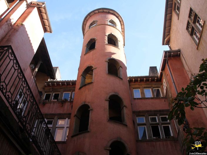 Une cour étroite à Lyon en France comprend une haute tour cylindrique de couleur pêche avec des fenêtres en arc ouvertes. Les bâtiments environnants sont de la même couleur avec de multiples fenêtres et toits, le tout sous un ciel bleu clair.