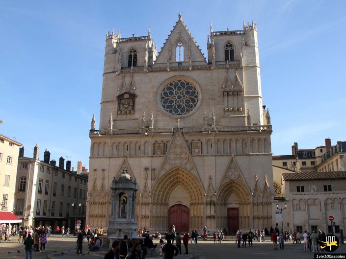Une grande cathédrale de style gothique avec une rosace et une façade complexe domine une place animée à Lyon en France. Des gens sont rassemblés devant, peut-être des touristes, sous un ciel bleu clair.