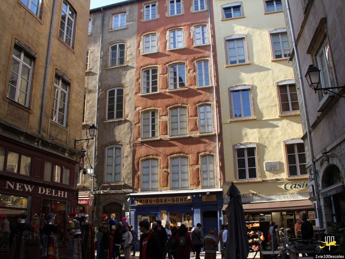 Une scène de rue dans une vieille ville européenne à Lyon en France avec des bâtiments colorés, divers magasins et des gens qui se promènent. Les bâtiments ont plusieurs fenêtres avec volets et la rue est très fréquentée par les piétons.