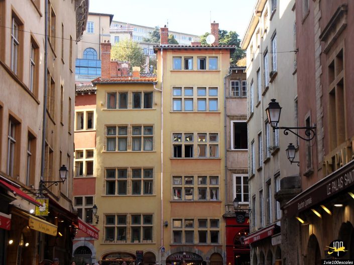 Une rue étroite à Lyon en France bordée de grands bâtiments historiques dotés de multiples fenêtres, balcons et devantures de magasins, avec un ciel clair au-dessus.
