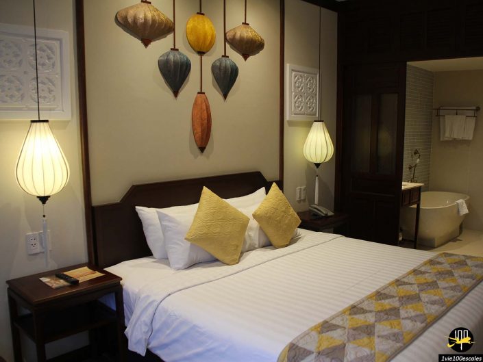 Une chambre bien éclairée avec un grand lit, une literie jaune et blanche, deux tables de chevet, des lanternes suspendues au dessus de la tête de lit et une baignoire visible dans une salle de bain attenante à Hoi An au Vietnam.