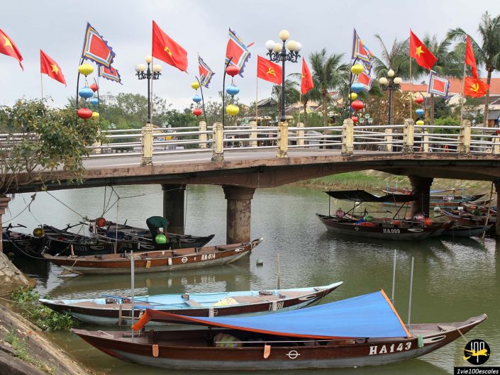 Une scène au bord de l'eau à Hoi An, au Vietnam, avec des bateaux traditionnels en bois amarrés sous un pont orné de drapeaux et de lanternes colorés. Des palmiers et des bâtiments sont visibles en arrière-plan.