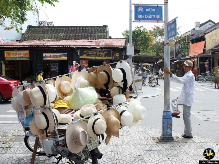 Un vélo chargé de divers chapeaux est garé au coin d’une rue. Un homme se tient à côté d'un poteau près des magasins et autres vélos de la rue Phan Chau Trinh à Hoi An au Vietnam.