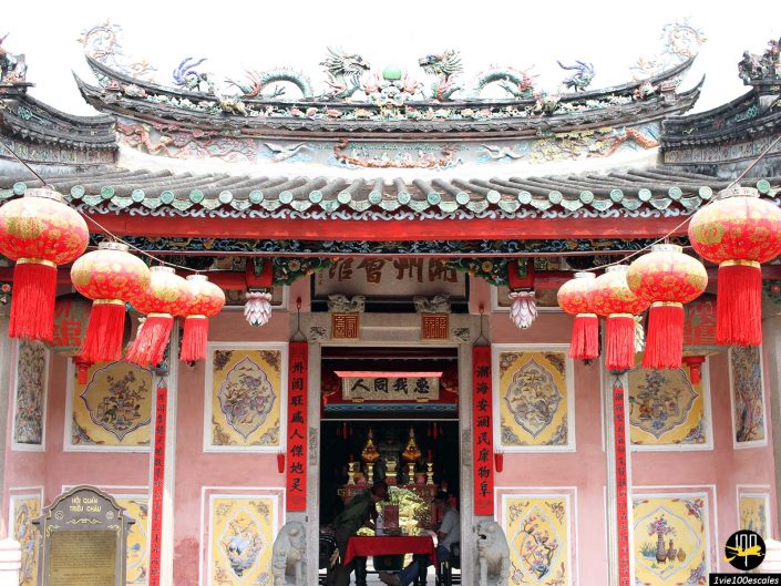 Vue de face de l'entrée d'un temple chinois traditionnel à Hoi An, au Vietnam, décorée de lanternes rouges et de sculptures complexes sur le toit. Des panneaux avec des caractères chinois sont accrochés au-dessus de la porte, offrant un aperçu de l'autel intérieur.