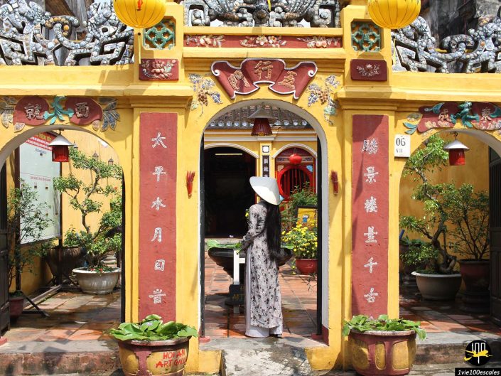 Une personne vêtue d'une tenue traditionnelle blanche et d'un chapeau conique se tient sous l'arche d'une passerelle jaune richement décorée avec des accents rouges et dorés et des caractères chinois à Hoi An au Vietnam.