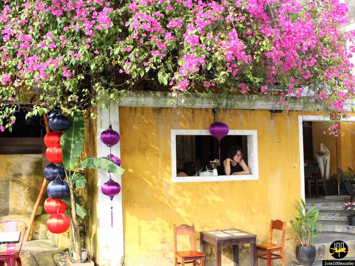 Une femme est assise à la fenêtre d'un bâtiment jaune à Hoi An au Vietnam, décoré de lanternes violettes et rouges au milieu de fleurs de bougainvilliers roses éclatantes. Deux chaises en bois et une petite table sont placées à l'extérieur.