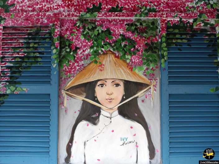 Une fresque murale représentant une femme portant un chapeau conique traditionnel portant une robe blanche, sur fond de volets bleus et de fleurs roses, évoque le charme d'Hoi An au Vietnam.