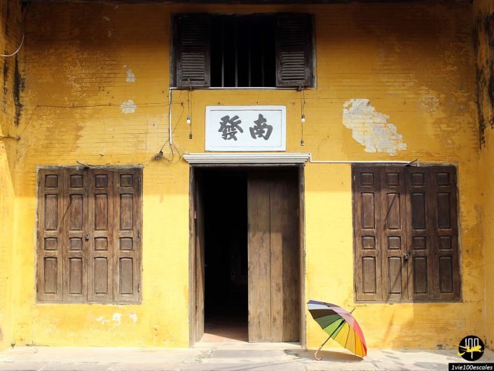 Un bâtiment jaune avec des fenêtres en bois et une porte ouverte vous accueille à Hoi An au Vietnam. Un parapluie multicolore est appuyé contre le mur près de l’entrée.