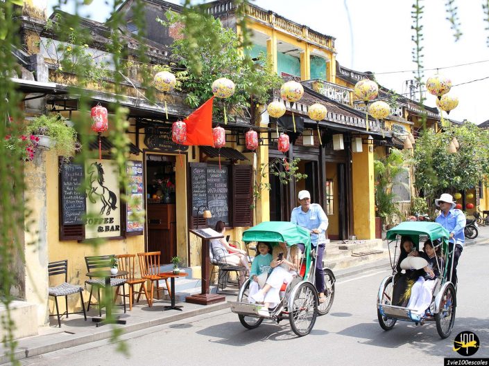 Des gens en cyclo-pousse dans une rue bordée de vieux bâtiments, de lanternes et d'un café-bar avec un coin salon extérieur à Hoi An au Vietnam.