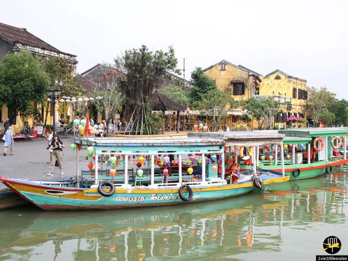 Des bateaux colorés sont amarrés le long d'une rivière urbaine à Hoi An au Vietnam, avec des passants à proximité. Des bâtiments traditionnels aux façades jaunes sont en arrière-plan.