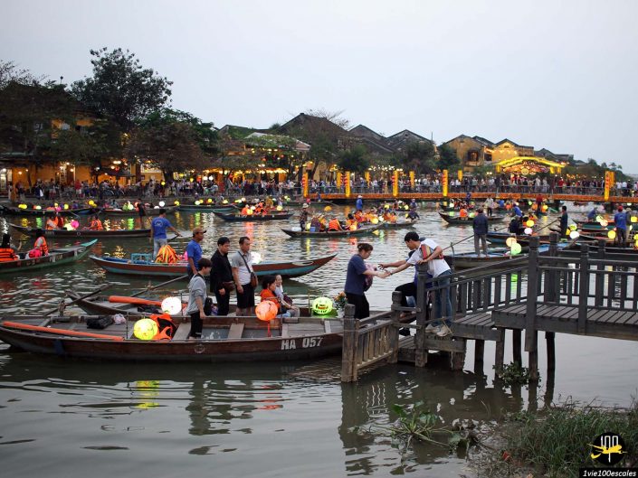 Une scène animée au bord d'une rivière au crépuscule à Hoi An, au Vietnam, avec de nombreuses personnes dans des bateaux éclairés par des lanternes et d'autres debout sur une jetée en bois. Des bâtiments et d’autres personnes sont visibles en arrière-plan.