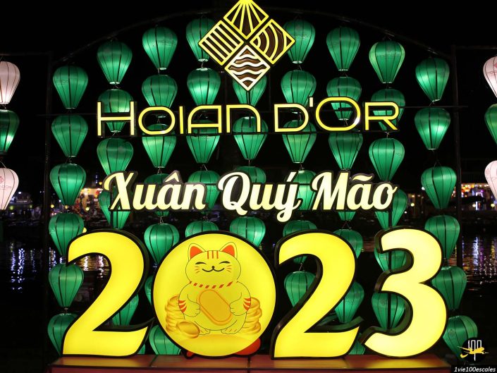 Un panneau lumineux décoratif indique « Hoi An D'Or », « Xuân Quý Mão » et « 2023 », avec des lanternes vertes et une figure centrale Maneki-neko, capturant l'essence vibrante de Hoi An au Vietnam.