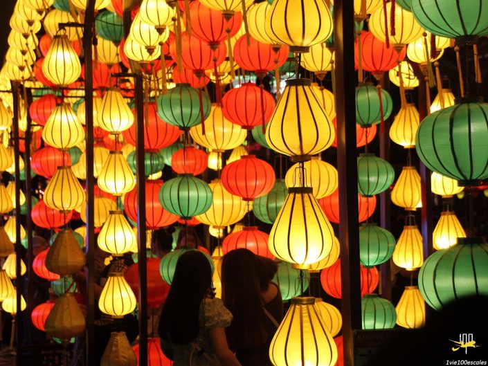 Une exposition vibrante de lanternes illuminées rouges, vertes et jaunes étroitement accrochées les unes aux autres à Hoi An au Vietnam, avec deux personnes debout en dessous.