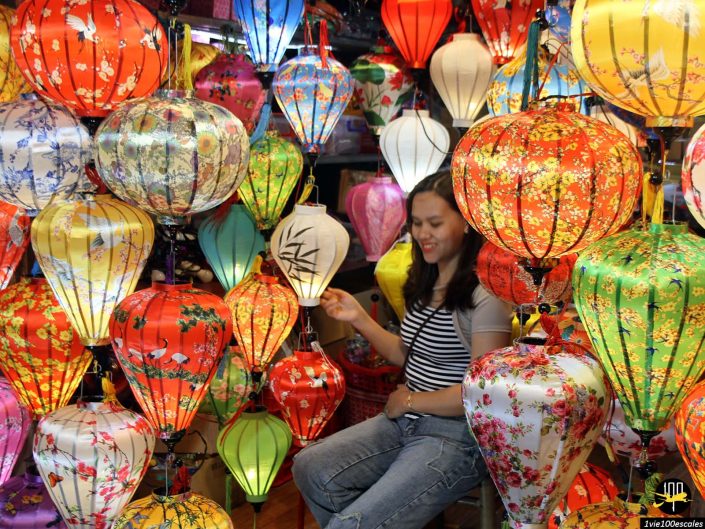 Une personne est assise parmi un éventail de lanternes suspendues colorées de différents motifs et designs dans un magasin à Hoi An au Vietnam.
