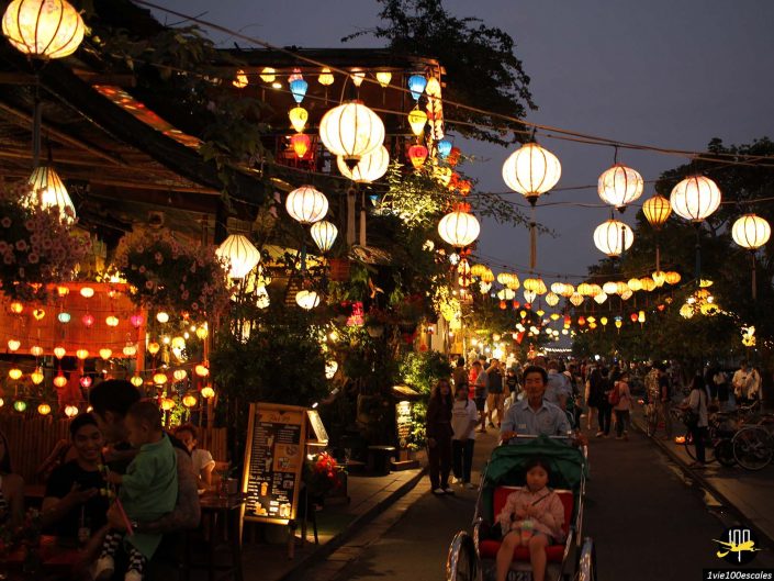Une rue animée à Hoi An au Vietnam au crépuscule, décorée de diverses lanternes colorées. Les gens marchent et un homme à vélo avec un enfant dans un pousse-pousse est au premier plan. Les magasins et les cafés sont éclairés sur les côtés.