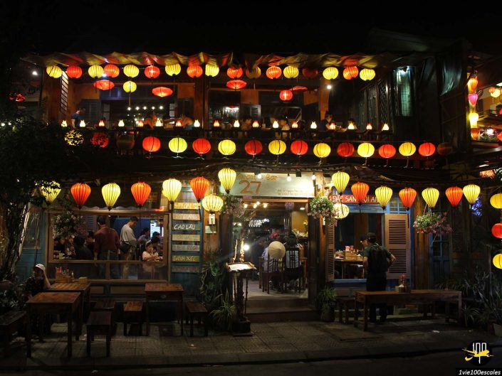 Un restaurant animé en bord de rue, à Hoi An au Vietnam, éclairé par de nombreuses lanternes colorées. Les convives sont assis à l’intérieur et au niveau supérieur, tandis que des tables vides sont visibles à l’extérieur.