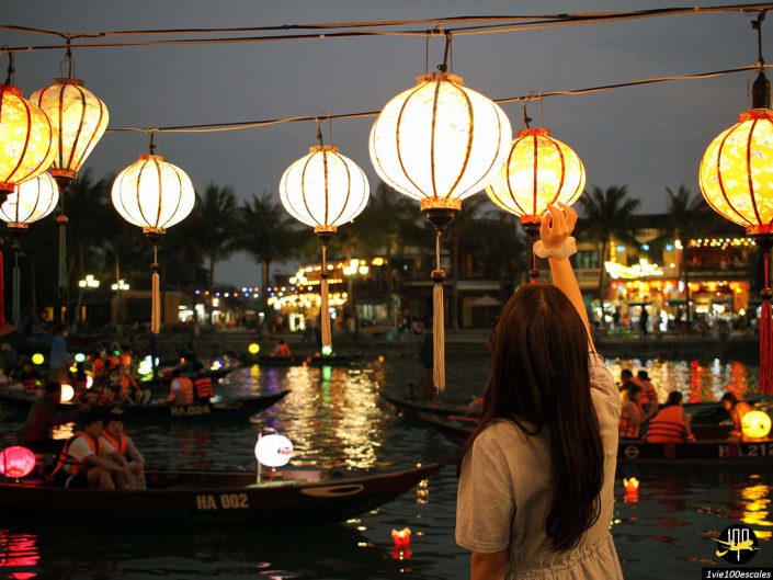 Une personne aux cheveux longs touche une lanterne sur un marché nocturne à Hoi An au Vietnam, avec des bateaux flottant dans l'eau ornés de lanternes et de lumières en arrière-plan.