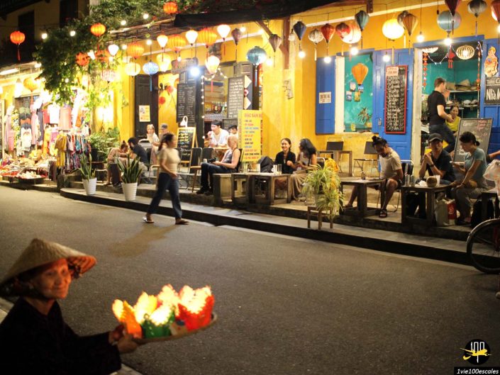 Une scène de rue nocturne animée à Hoi An au Vietnam avec des gens dînant dans des restaurants en plein air près d'un bâtiment jaune bien éclairé. Une personne portant un chapeau conique porte un plateau de lanternes en papier allumées, tandis que des lanternes colorées sont suspendues au-dessus de sa tête.