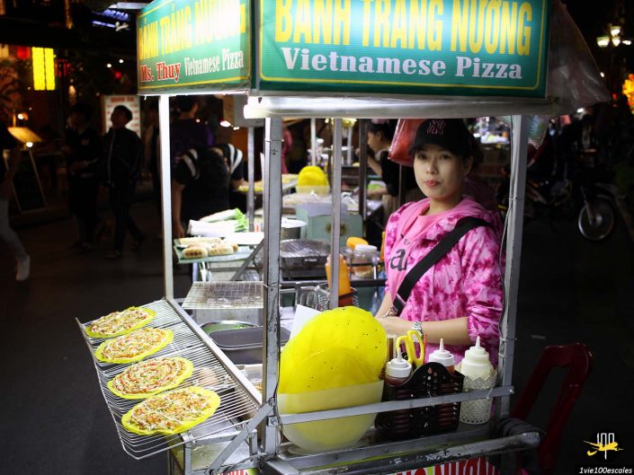 Un vendeur ambulant en tenue rose se tient derrière un chariot étiqueté « Pizza vietnamienne » à Hoi An au Vietnam, avec plusieurs pizzas exposées sur des grilles. Un gros objet jaune et des condiments sont également visibles sur le chariot.