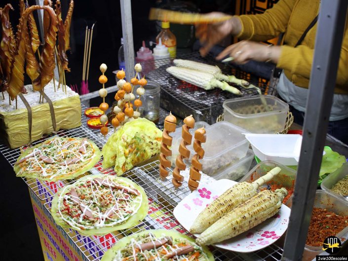 Un vendeur de nourriture de rue à Hoi An, au Vietnam, prépare et sert divers aliments grillés et en brochettes, notamment des épis de maïs, des brochettes coupées en spirale et des pains plats garnis de sauces et de garnitures.