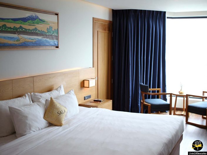 Une chambre d'hôtel bien éclairée avec un grand lit, des draps blancs, une tête de lit en bois, un tableau au-dessus du lit et un coin salon avec deux chaises et une petite table près d'une fenêtre aux rideaux sombres à Da Nang au Vietnam.