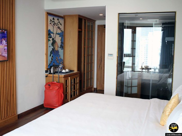 Une chambre d'hôtel avec un lit soigneusement fait, un sac à dos rouge sur un porte-bagages, une télévision fixée au mur, un petit coin kitchenette et une fenêtre en verre montrant la vue animée sur la ville de Da Nang au Vietnam à l'extérieur.