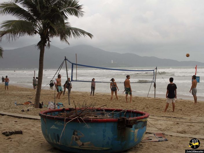 Des gens jouant au beach-volley près de l'océan avec des montagnes en arrière-plan à Da Nang au Vietnam. Une cuve en métal bleu et délabrée avec des images peintes se trouve au premier plan, à côté d’un palmier.