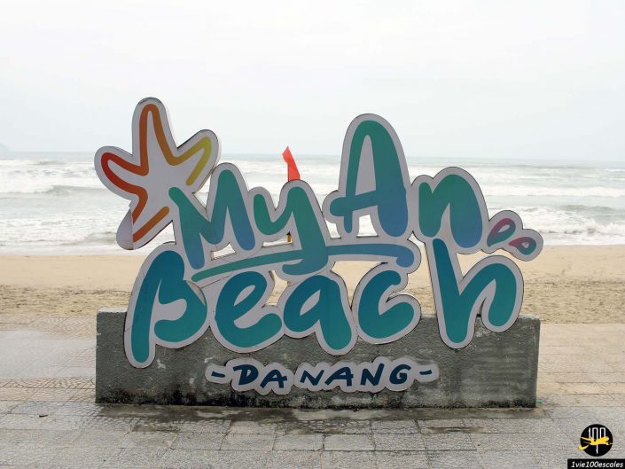 Un panneau indiquant « My An Beach Da Nang » en lettres colorées est placé devant une plage de sable avec des vagues s'écrasant en arrière-plan, à Da Nang au Vietnam.