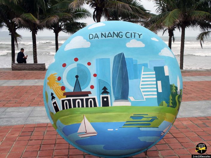 Une grande sculpture sphérique présente des monuments illustrés de la ville de Da Nang à Da Nang au Vietnam, situés sur une promenade en bord de mer avec des palmiers, une vue sur l'océan et une personne assise sur un banc en arrière-plan.