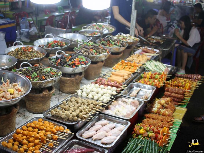 Un étal de marché à Da Nang au Vietnam présente une gamme de produits alimentaires de rue, notamment des brochettes, des légumes grillés et des plats dans des bols en métal. Les gens sont visibles en arrière-plan.