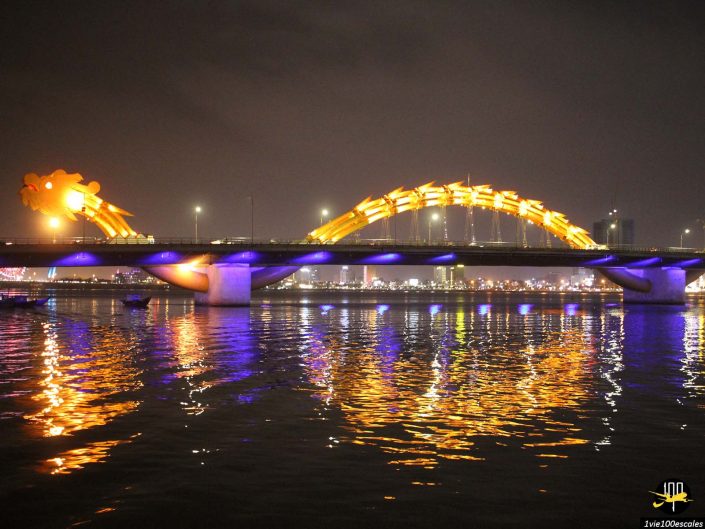 Un pont conçu pour ressembler à un dragon, éclairé par des lumières jaunes et violettes, enjambe un plan d'eau la nuit à Da Nang, au Vietnam, avec les lumières de la ville visibles en arrière-plan.