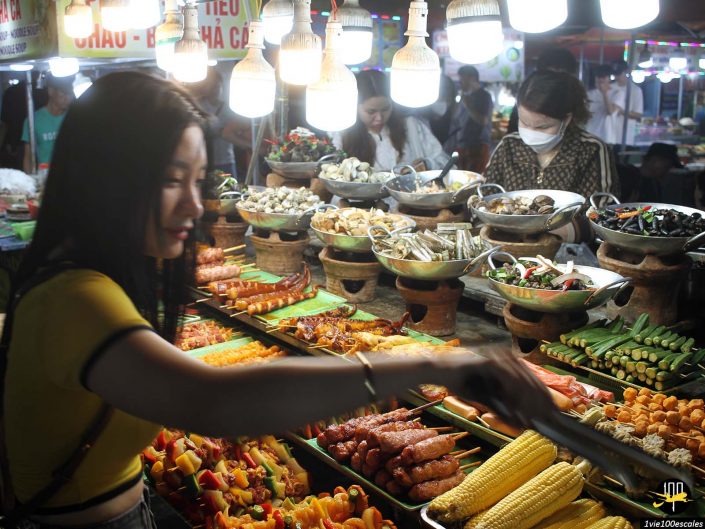 Des gens sélectionnent et se servent divers aliments dans un stand de marché de rue à Da Nang au Vietnam, avec un éclairage lumineux illuminant l'assortiment diversifié de légumes, de viandes et de fruits de mer.