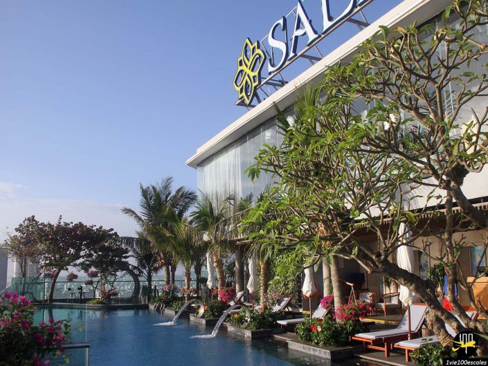 Espace piscine sur le toit avec chaises longues, plantes tropicales et jeux d'eau en cascade dans un hôtel à Da Nang au Vietnam, avec signalétique "SALA" visible sur le bâtiment.