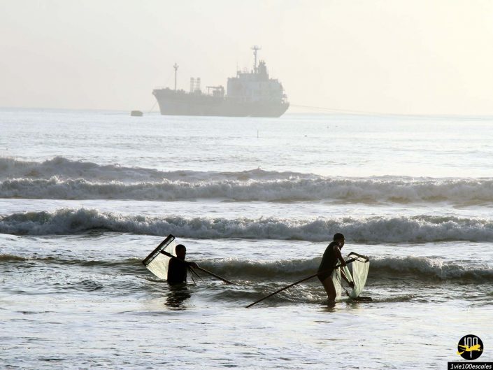 Deux personnes pataugent dans les eaux peu profondes de l'océan, tenant de grands filets de pêche, avec un grand navire en arrière-plan, par une journée brumeuse à Da Nang au Vietnam.
