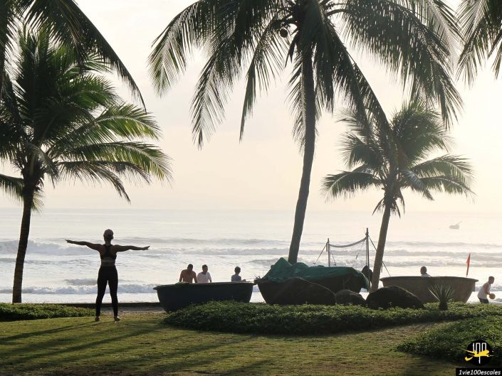 Une personne s'étend sur une zone herbeuse près de la plage à Da Nang au Vietnam, avec plusieurs personnes dans ou près de l'eau et des bateaux sous les palmiers en arrière-plan.
